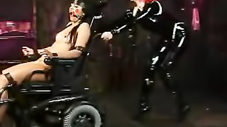 Naked hottie gets pinned down by metal pipes on a wooden floor then gets her pussy whipped by a redhead chick in black latex outfit before she gets tied on a black wheel chair and gets gagged by a red ball then gets stepped on her mouth.
