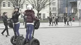 Two sexy brunette girls ride segways in park square while wearing coats.